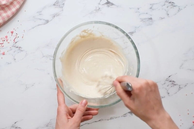stirring melted white chocolate in a glass bowl with a whisk.