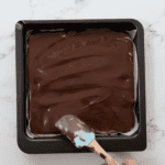 spreading chocolate over peanut butter in a baking dish with a rubber spatula.