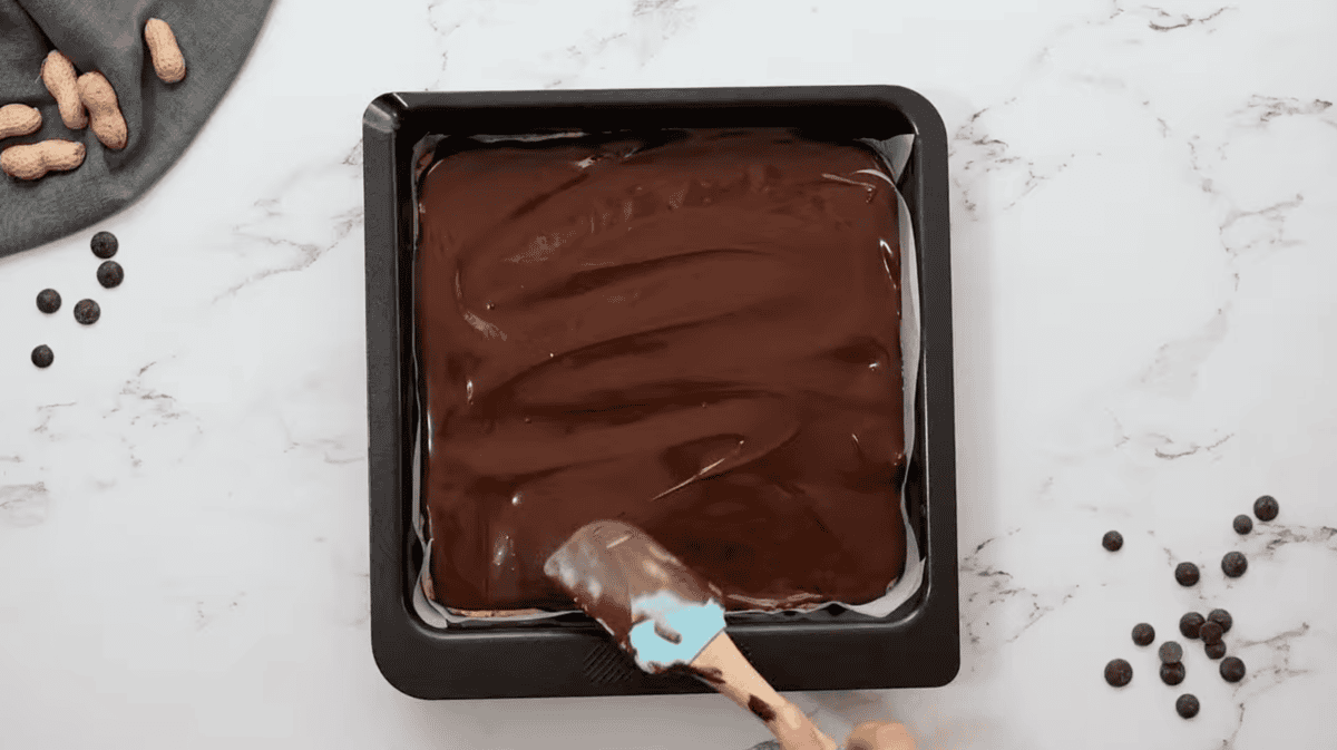 spreading chocolate over peanut butter in a baking dish with a rubber spatula.