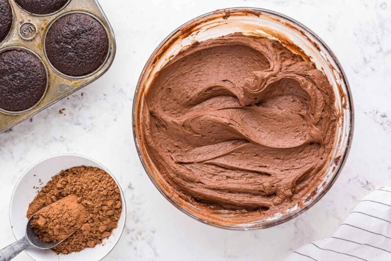 chocolate buttercream frosting in a bowl next to chocolate cupcakes and cocoa powder.