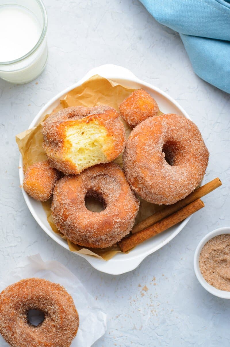 bitten donut on top of cinnamon sugar donuts on a white serving dish with cinnamon sticks.