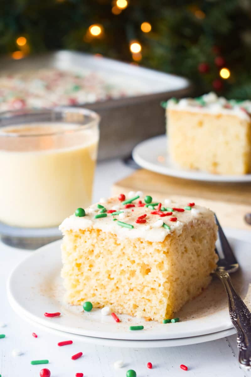 a slice of eggnog sheet cake on a white plate with a fork and a glass of eggnog.