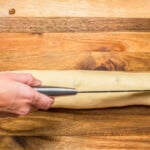 chefs knife cutting rolled filled estonian kringle dough in half lengthwise on a wood cutting board.