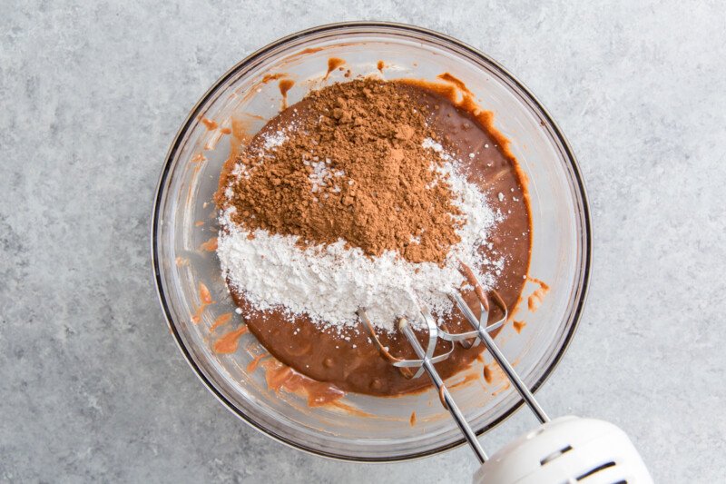 dry ingredients added to fudgy brownie batter in a glass bowl with a hand mixer.