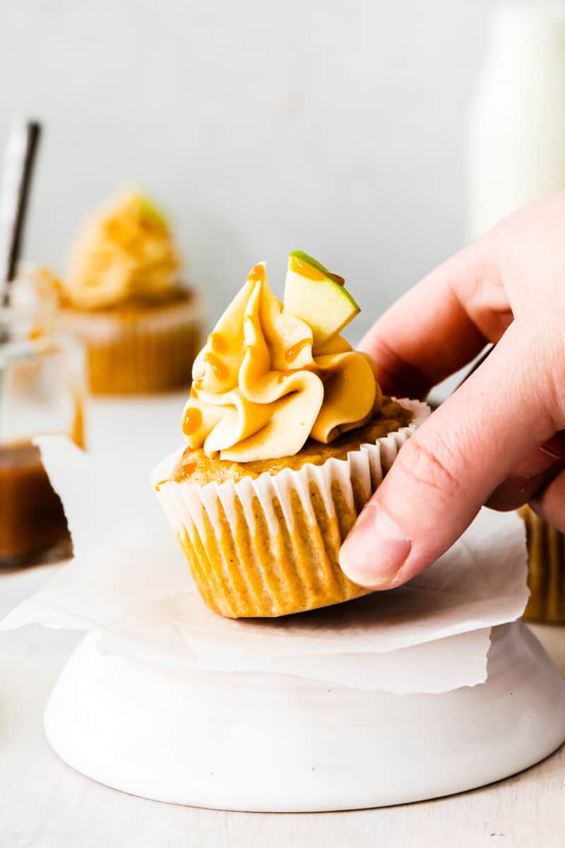 hand holding apple cupcake topped with caramel frosting, a drizzle of salted caramel, and an apple slice