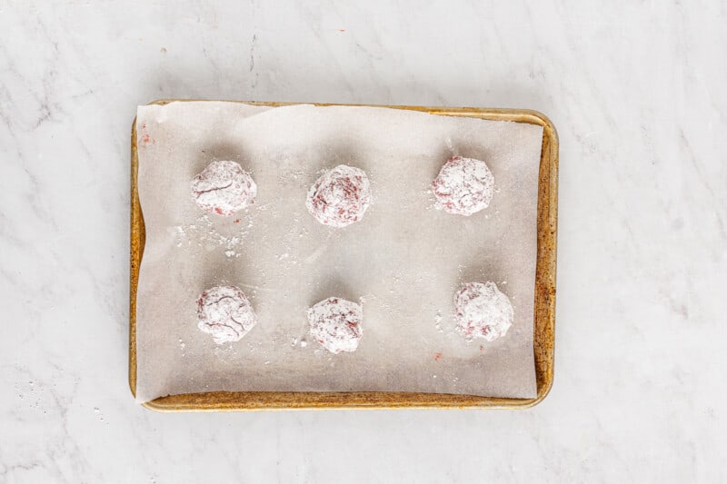 6 cheesecake stuffed cookie dough balls rolled in powdered sugar on a baking sheet lined with parchment paper