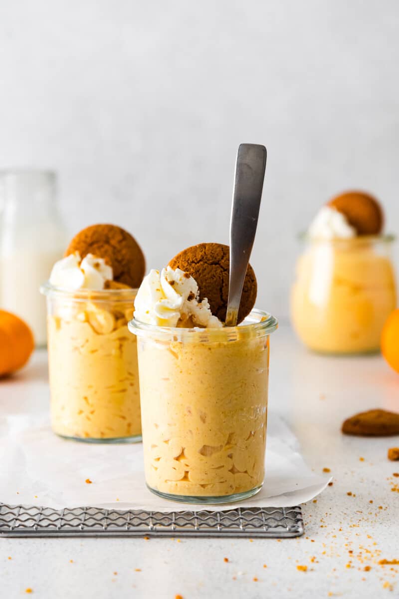 2 glass jars of pumpkin mousse topped with whipped cream and gingersnap cookies with a spoon in one jar