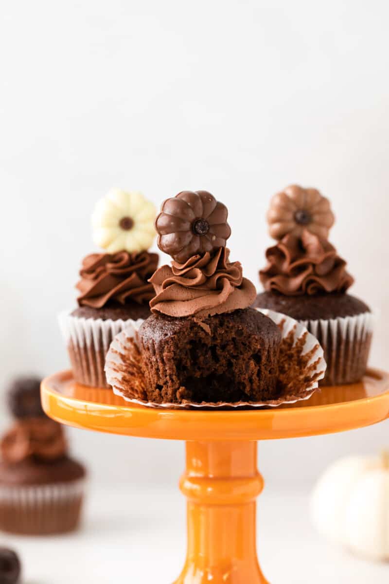 3 chocolate pumpkin cupcakes and one unwrapped cupcake with a bite taken out on an orange cake stand.