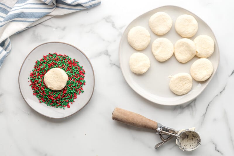 flattened sugar cookie dough balls on a white plate with one cookie dough ball in a plate of red and green sprinkles