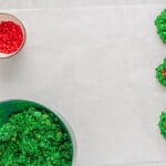 cornflake wreath mixture and red candies in bowls with 3 cornflake wreaths on parchment paper