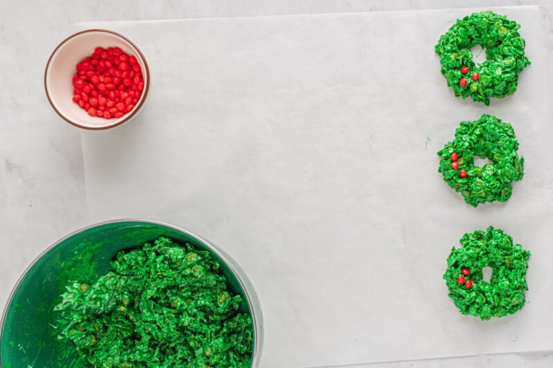 cornflake wreath mixture and red candies in bowls with 3 cornflake wreaths on parchment paper