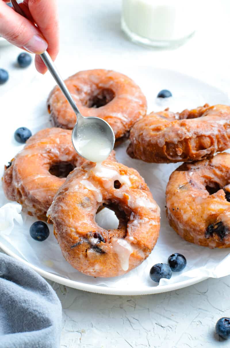 hand using a spoon to drizzle glaze onto blueberry donuts on a white plate
