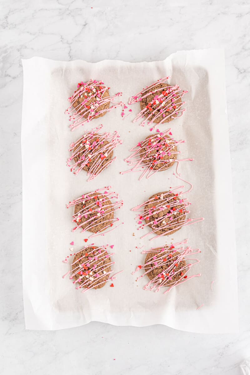 chocolate sugar cookies decorated with a drizzle of pink icing and Valentine's Day sprinkles on a parchment paper lined baking sheet