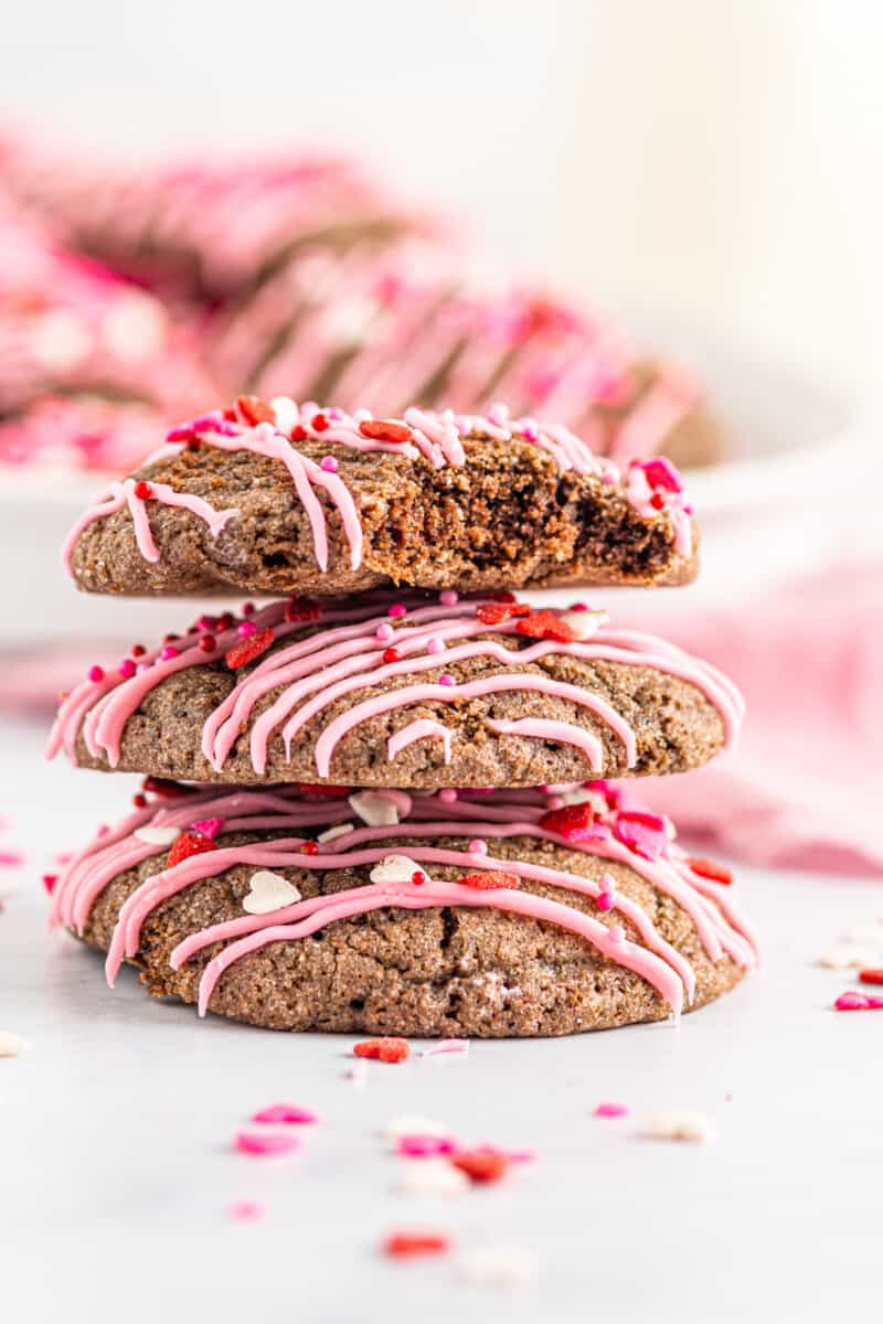 stack of 3 chocolate sugar cookies decorated with a drizzle of pink icing and Valentine's Day sprinkles with a bite taken from one