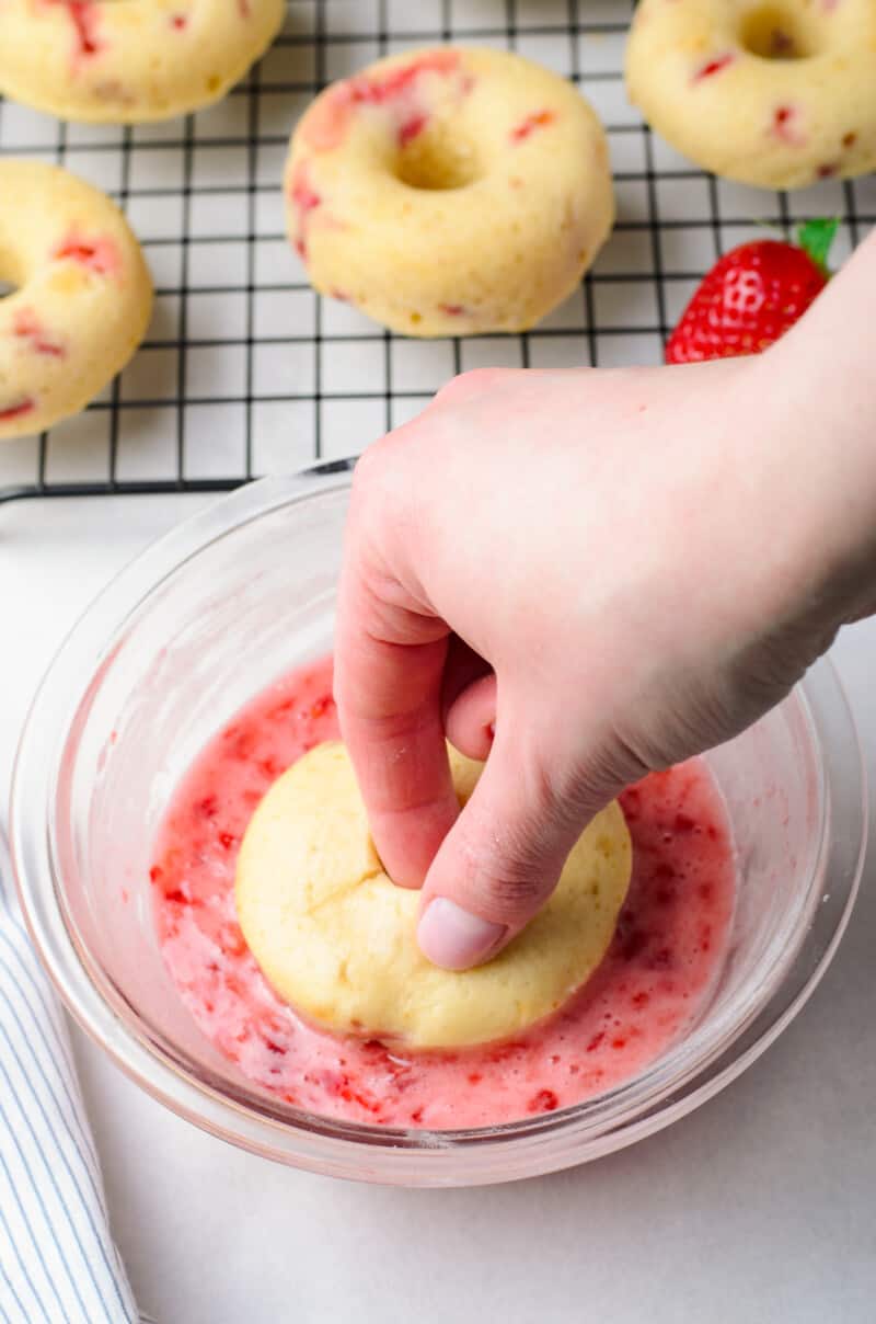 hand dipping a strawberry donut into strawberry glaze in a glass bowl