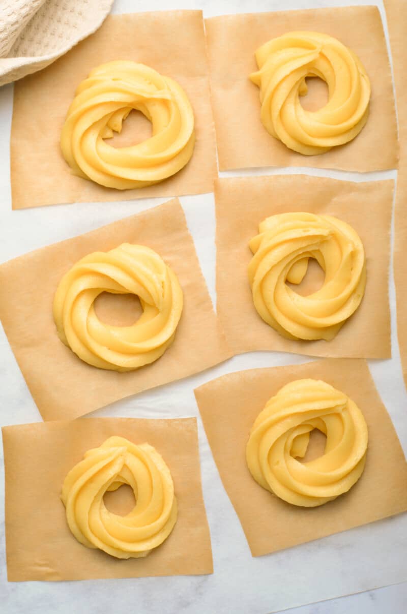 piped cruller dough in circles on squares of parchment paper