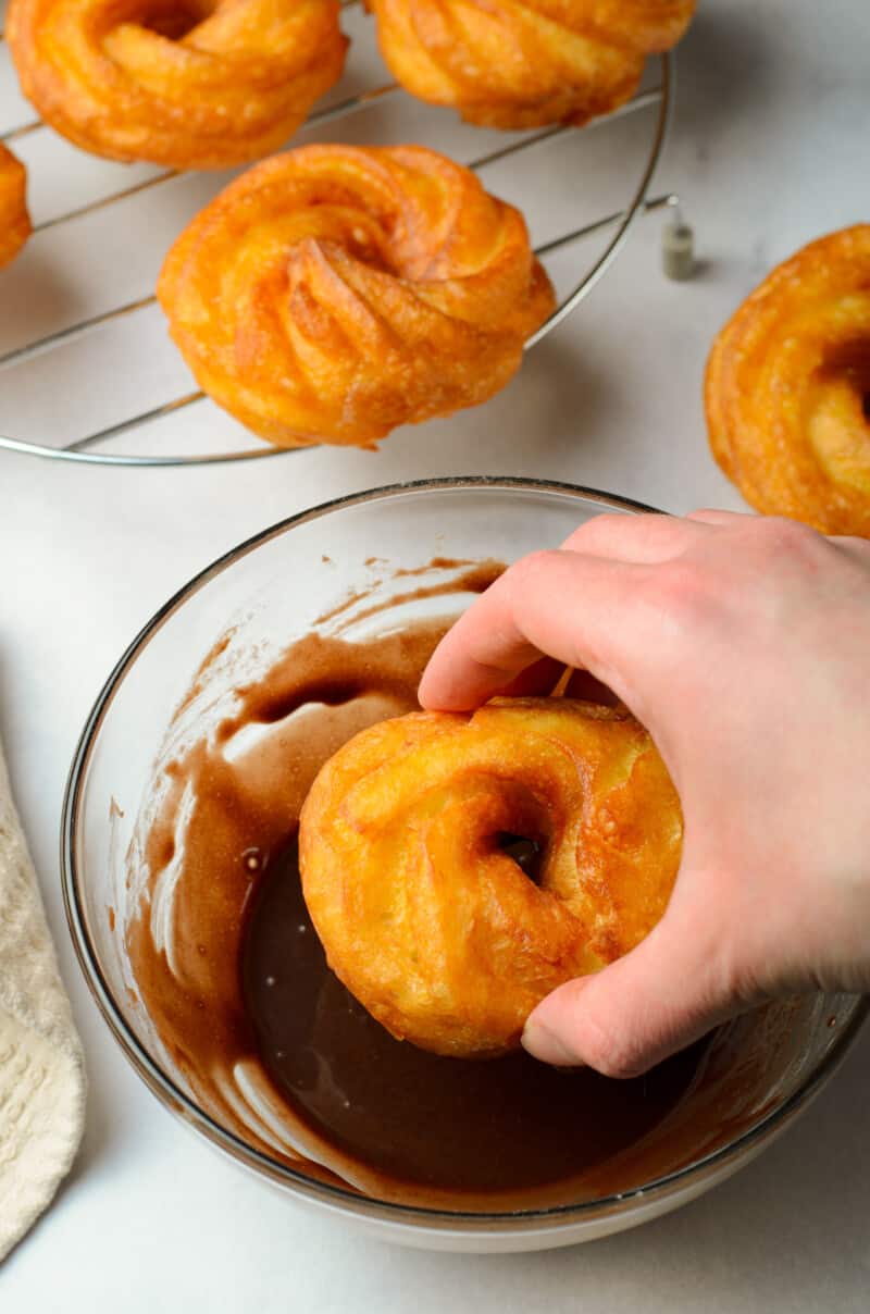 hand dipping a cruller into a bowl of chocolate glaze