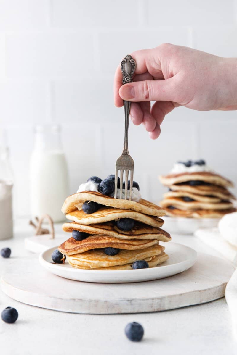 stack of blueberry pancakes on a white plate with a hand with a fork