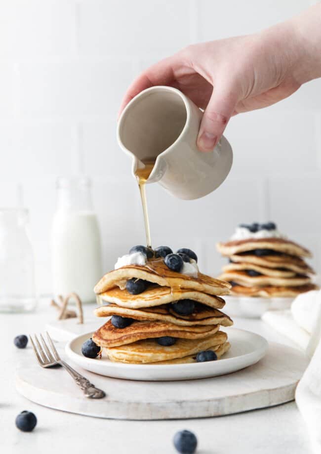 hand pouring maple syrup from a white container onto a stack of blueberry pancakes on a white plate