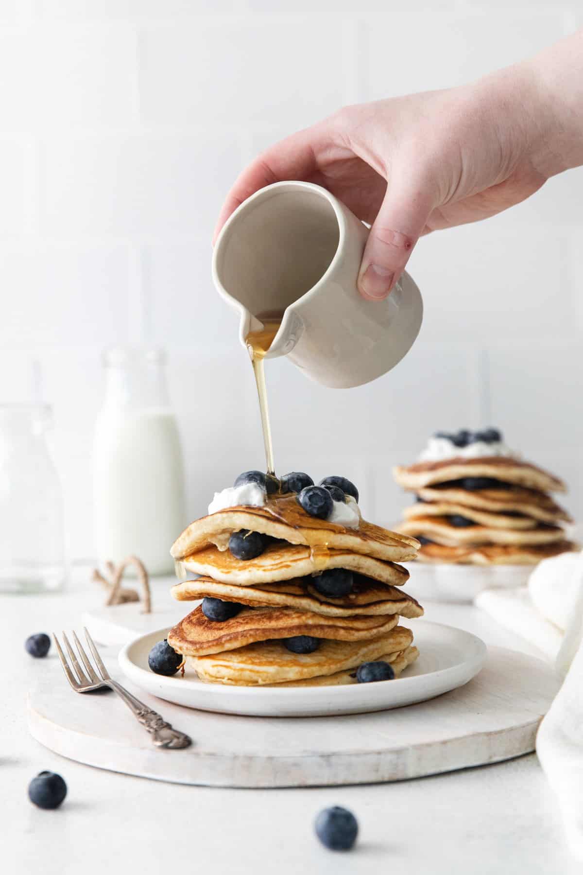 hand pouring maple syrup from a white container onto a stack of blueberry pancakes on a white plate