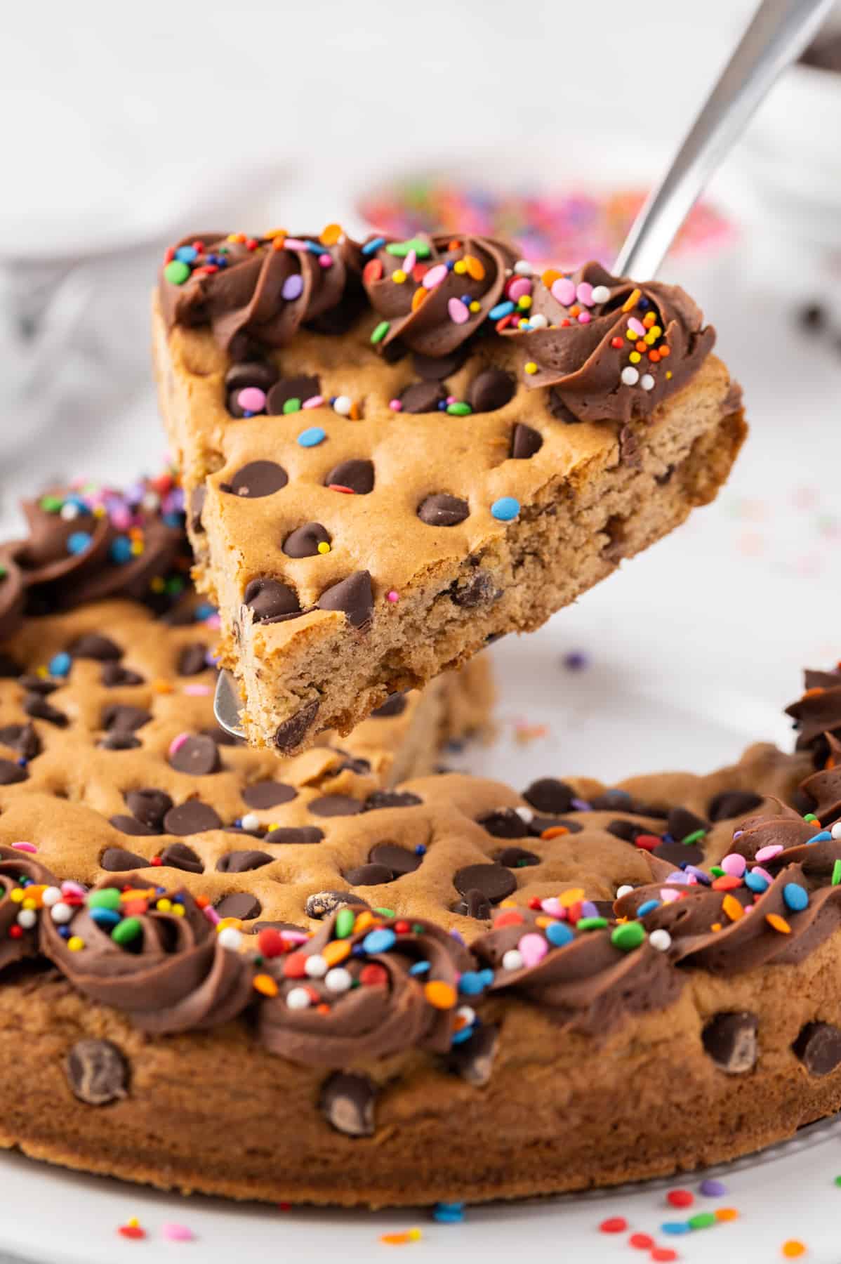 slice of chocolate chip cookie cake with chocolate frosting and sprinkles on a spatula