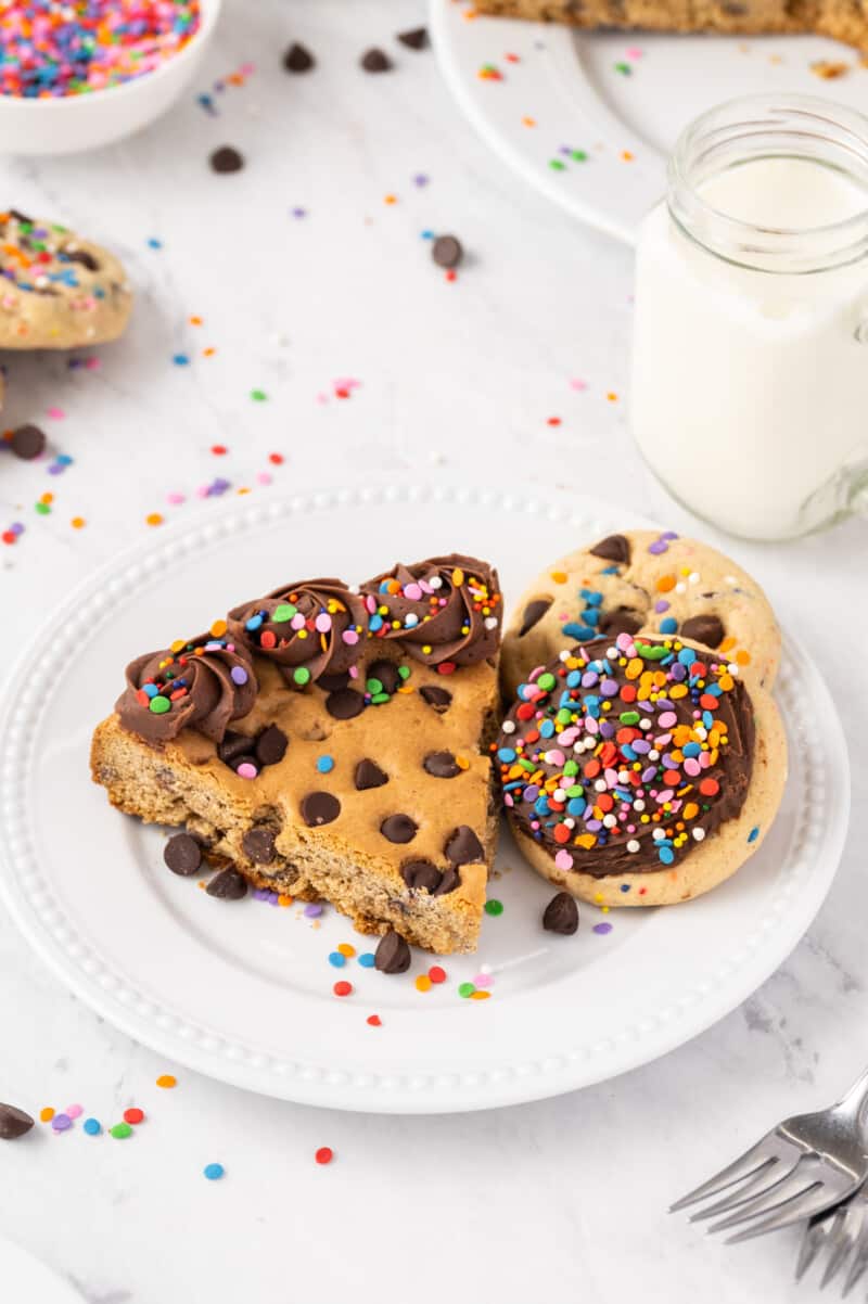 slice of chocolate chip cookie cake with chocolate frosting and sprinkles on a white plate with cookies