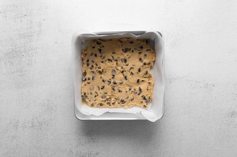 chocolate chip cookie dough in the bottom of a square parchment paper lined baking pan