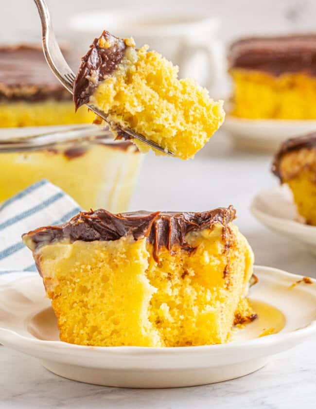 a forkful of boston cream pie poke cake suspended above a slice on a white plate.