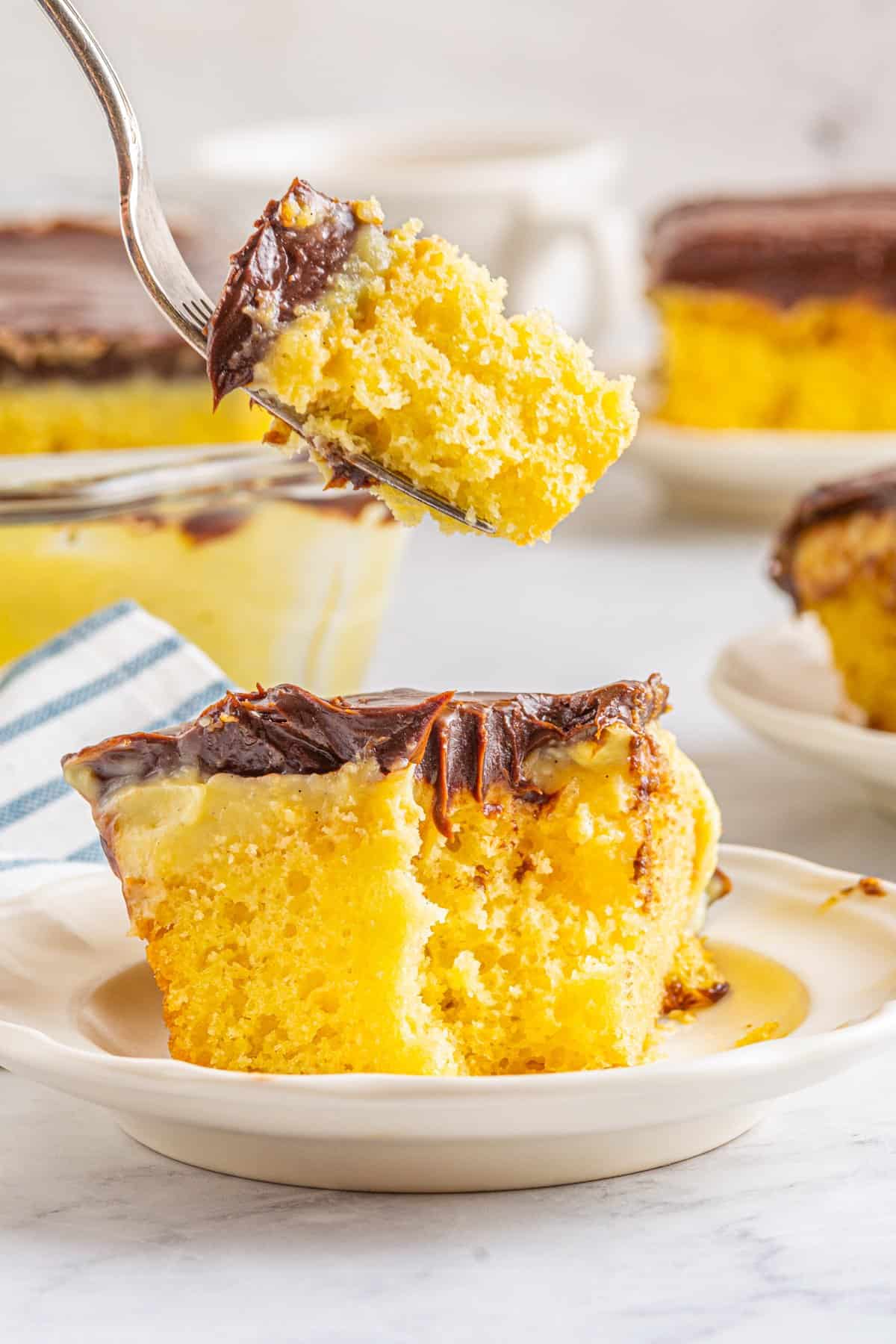 a forkful of boston cream pie poke cake suspended above a slice on a white plate.