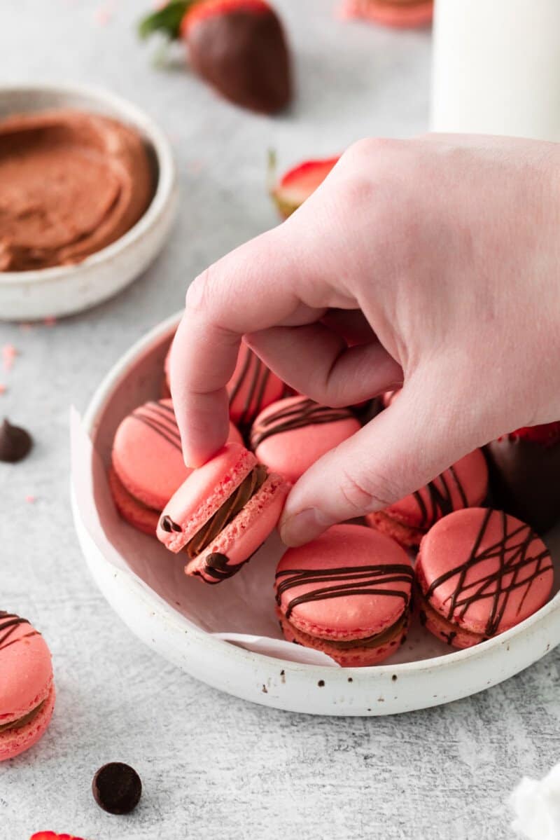 a hand grabbing a chocolate strawberry macaron from a white bowl.