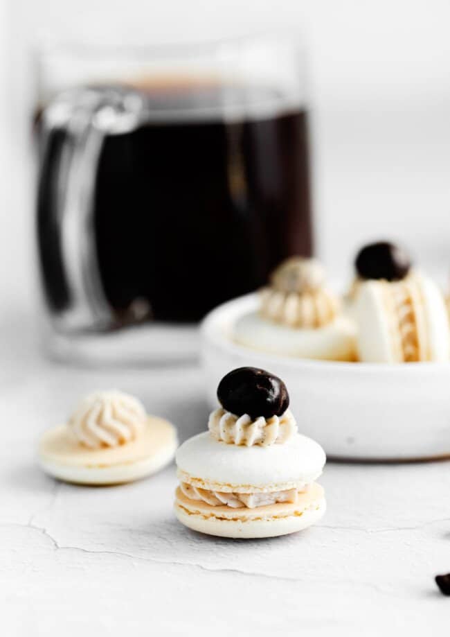 close up of a coffee macaron in front of a white bowl of coffee macarons.