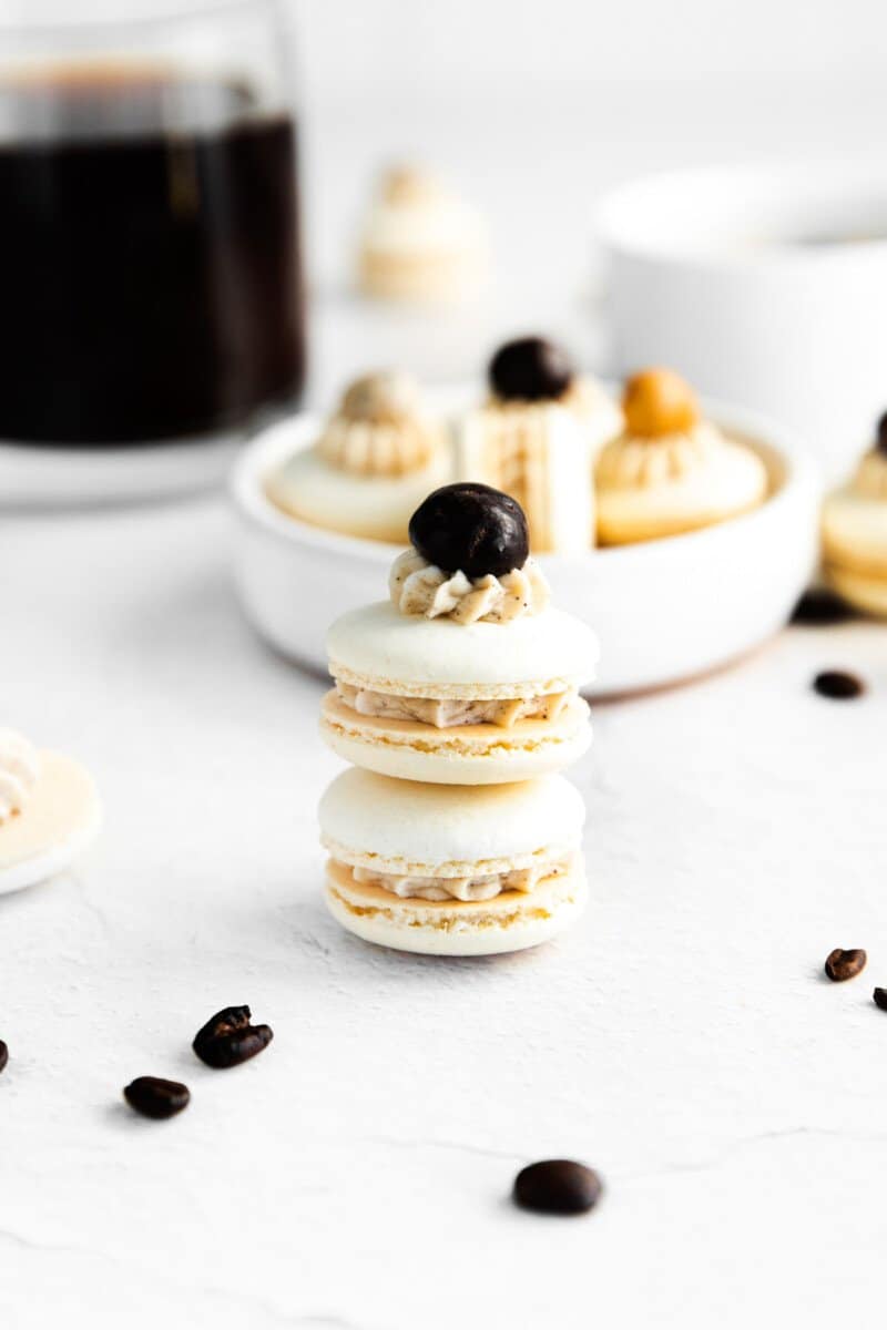 2 coffee macarons stacked in front of a white bowl of macarons.