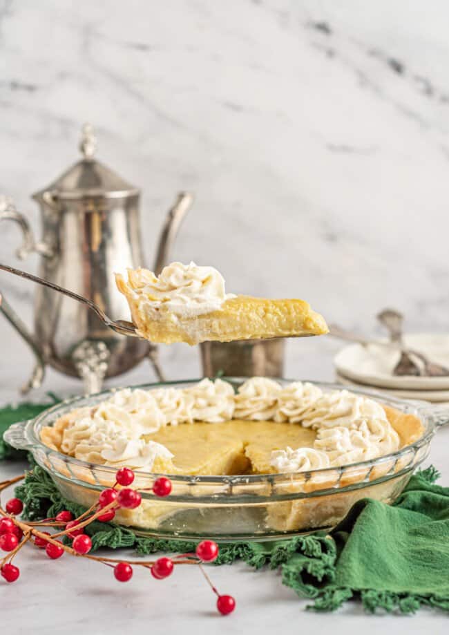 a slice of eggnog pie on a pie server hovering above eggnog pie in a glass pie dish.