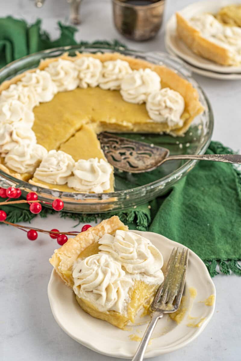 a slice of eggnog pie on a white plate with a fork in front of a sliced eggnog pie in a glass pie dish.