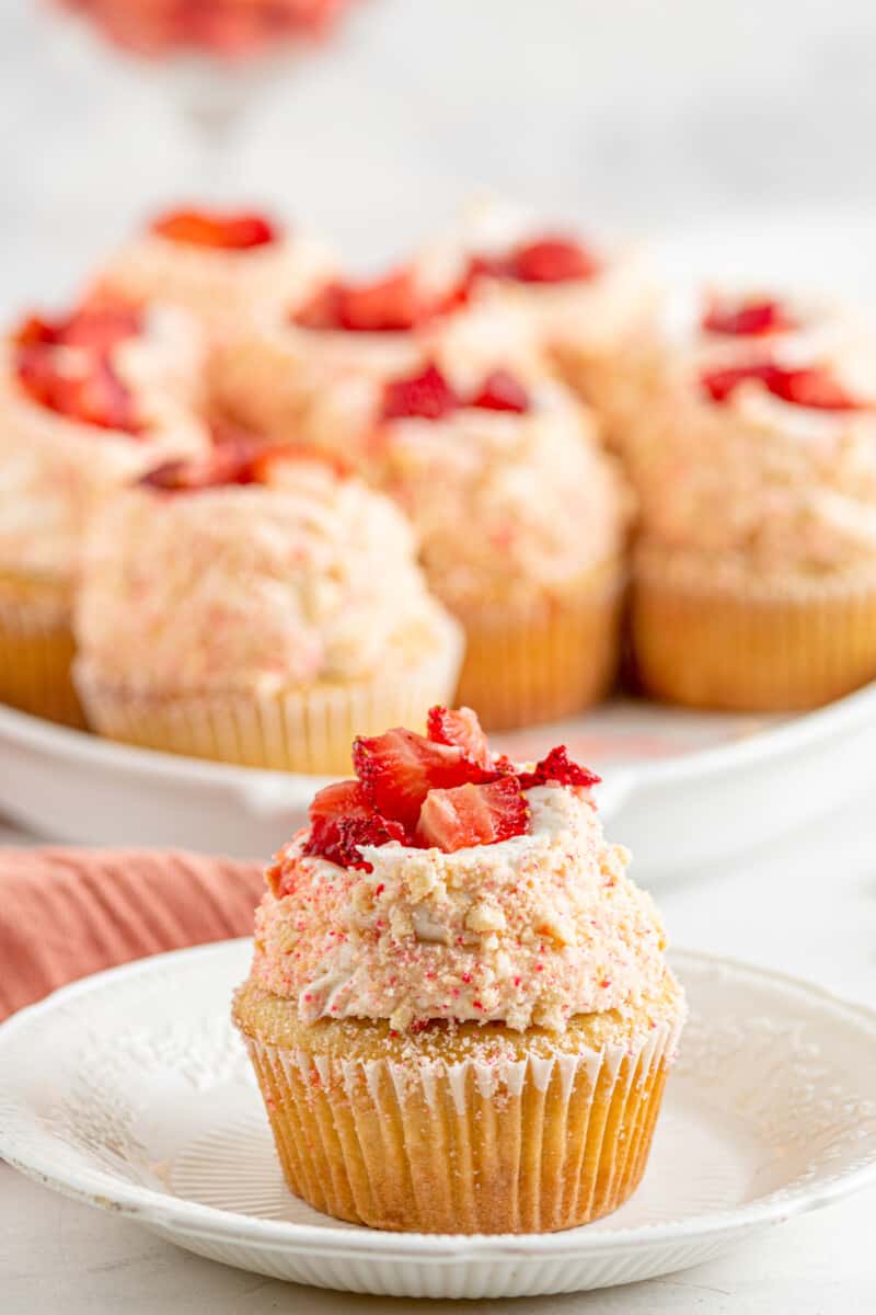 strawberry shortcake cupcake on a white plate in front of a tray of cupcakes.