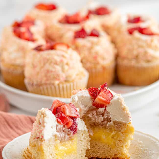 bisected strawberry shortcake cupcake on a white plate in front of a tray of cupcakes.