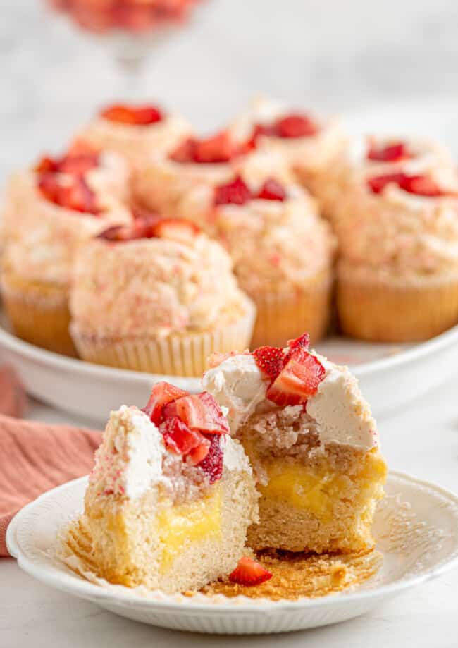 bisected strawberry shortcake cupcake on a white plate in front of a tray of cupcakes.