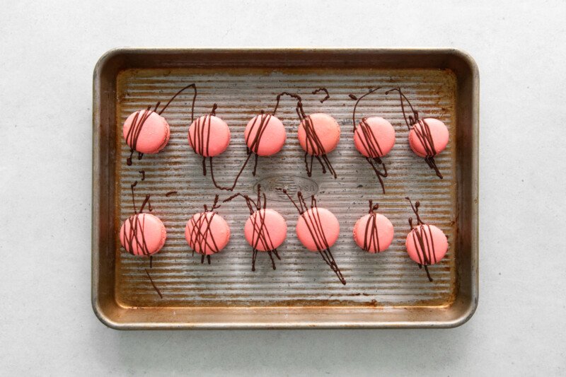 overhead view of 12 chocolate drizzled chocolate strawberry macarons on a baking sheet.