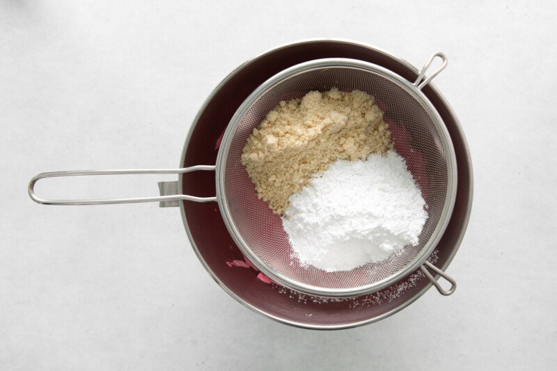 overhead view of sugar and almond flour sifted into pink meringue in a stainless mixing bowl.