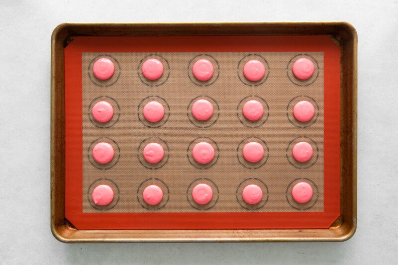 overhead view of 20 piped pink macaron shells on a silpat lined baking sheet.