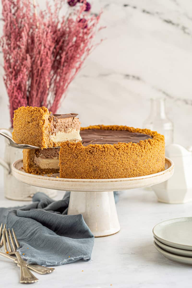 a slice of chocolate caramel cheesecake being lifted off of a white cake stand by a cake server.