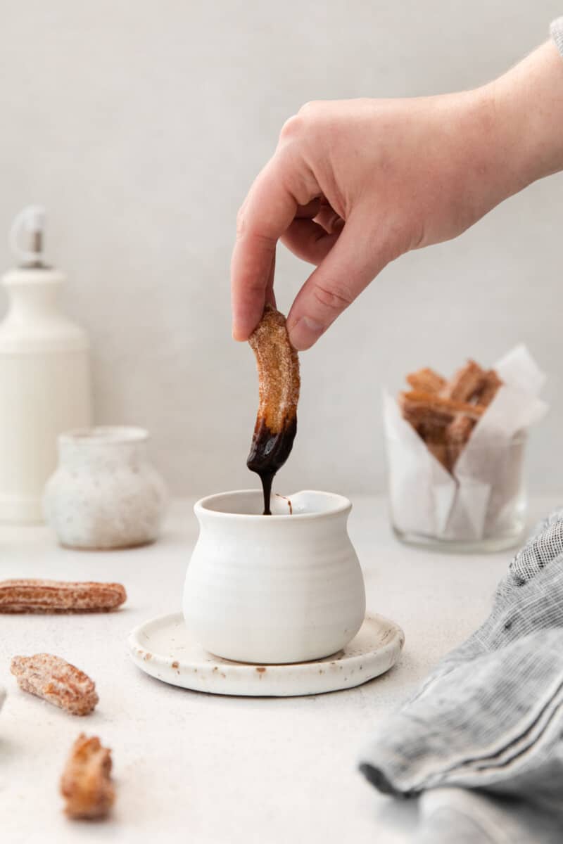 hand dipping a churro into chocolate sauce in a white pot.