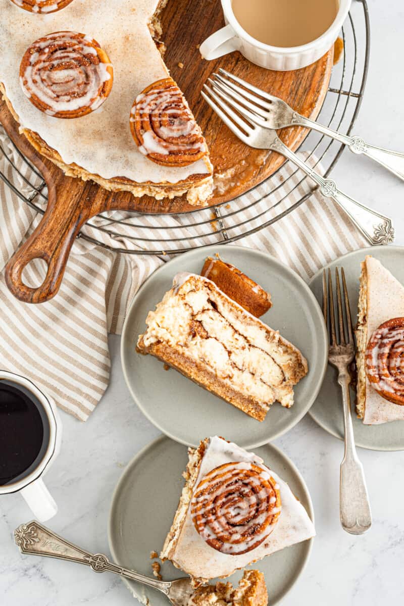 slices of cinnamon roll cheesecake on gray plates next to a sliced cinnamon roll cheesecake on a wooden platter.