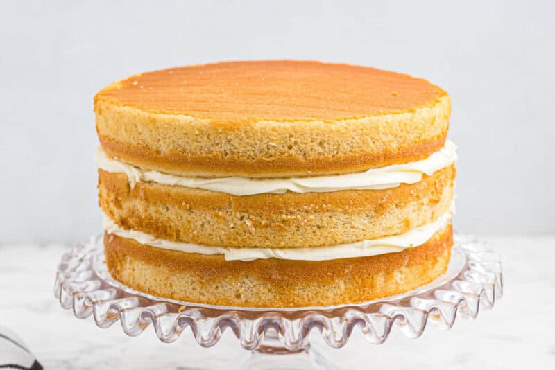 3 stacked wedding cake layers with buttercream frosting between them on a glass cake stand.