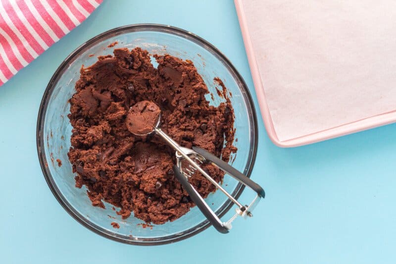 a cookie scoop full of chocolate pudding cookie dough in a glass bowl full of chocolate pudding cookie dough.