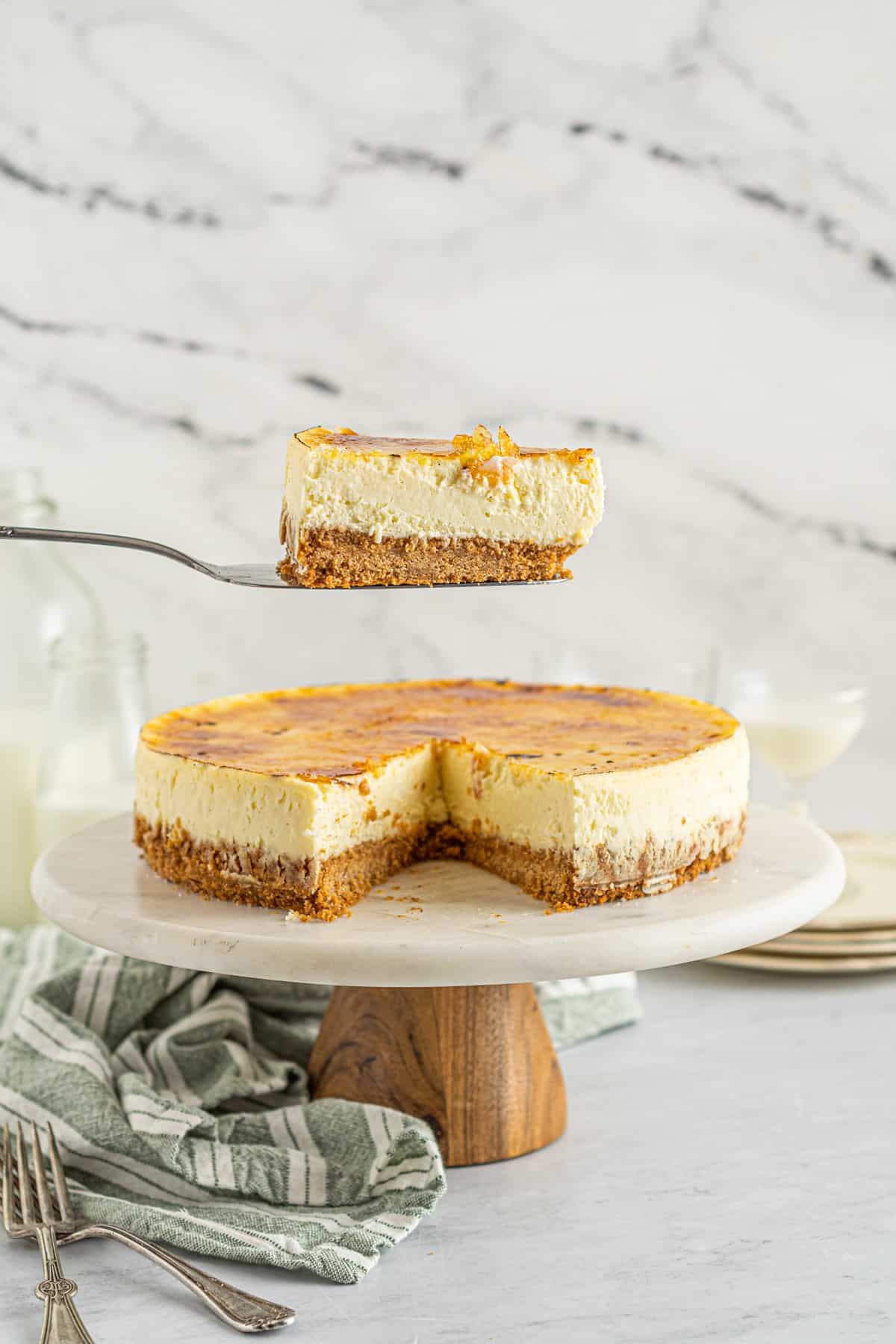 a slice of creme brulee cheesecake on a cake server hovering over a sliced cheesecake on a white cake stand with a wooden base.