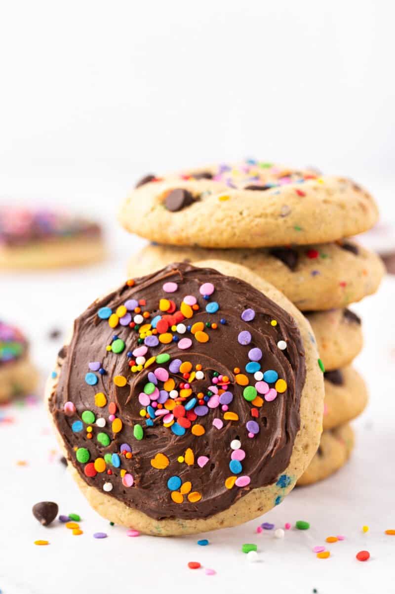 a frosted chocolate chip cookie leaning against a stack of 5 chocolate chip cookies.