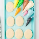 overhead view of 6 easter egg sugar cookies on a blue baking sheet with piping bags filled with colored frosting.