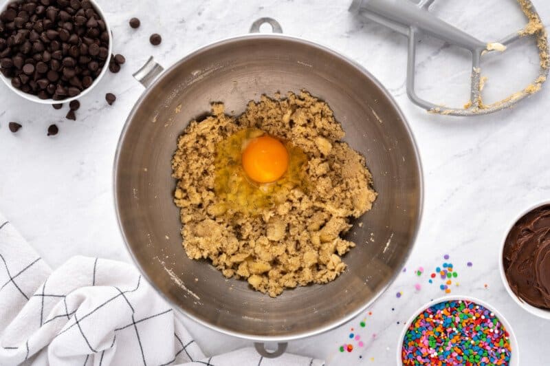 overhead view of egg added to frosted chocolate chip cookie dough in a stainless mixing bowl.