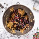overhead view of chocolate chips and sprinkles added to frosted chocolate chip cookie dough in a stainless mixing bowl.
